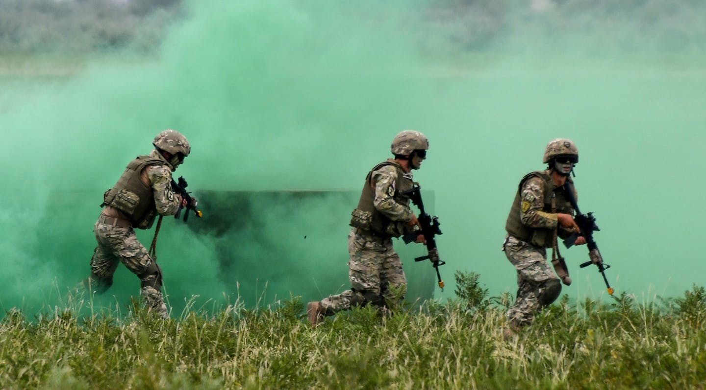 Three military members in camouflage and wearing helmets move through turf holding guns
