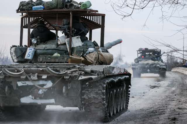 Two dark green tanks, as seen from behind, moving along a snowy road.