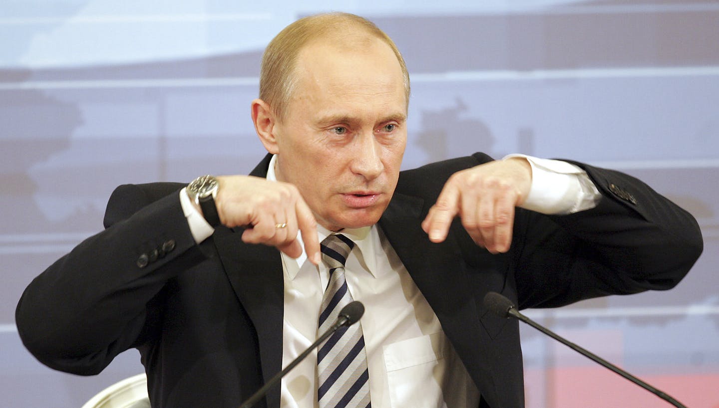 Vladimir Putin in a dark jacket and white shirt speaking into a microphone while gesturing with his hands.