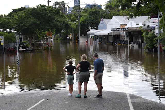 Three people stand on the edge of floodwaters in Queensland.
