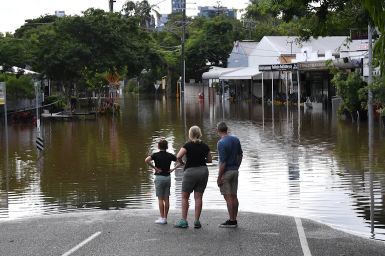 Three people stand on the edge of floodwaters in Queensland.