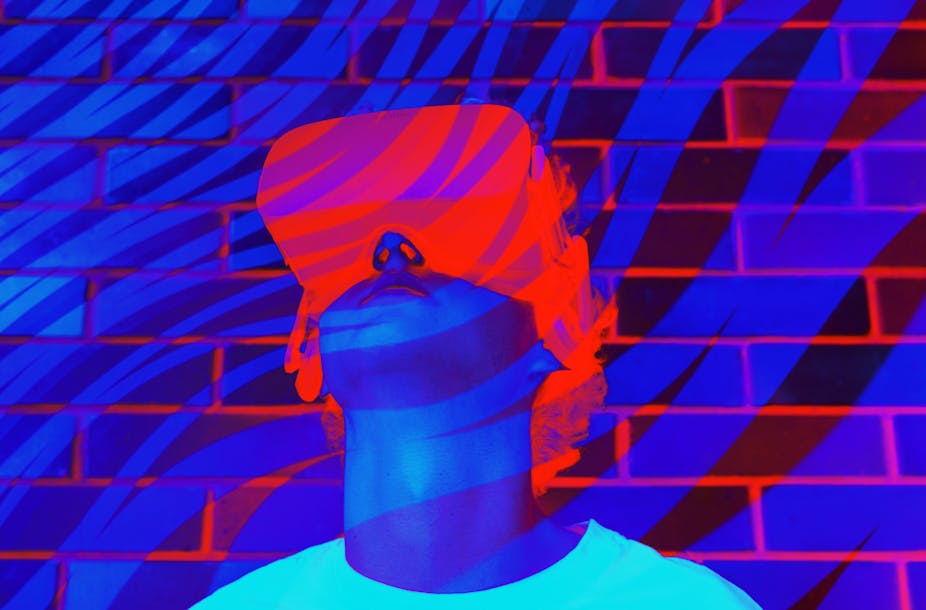 A young man or teenager in a white t-shirt wears a virtual reality headset with fluorescent blue, purple and orange colors in the background.