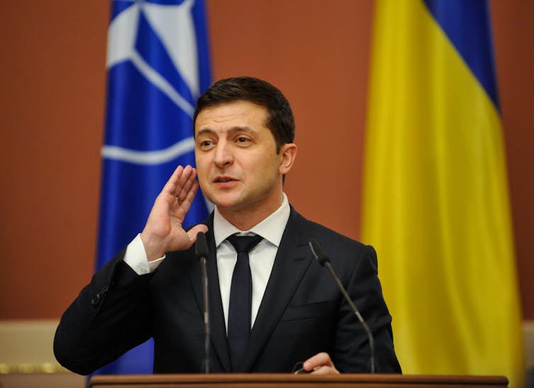 A man in a black suit stands behind a lectern with flags in the background.
