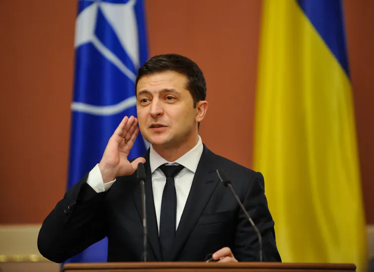 A man in a black suit stands behind a lectern with flags in the background.