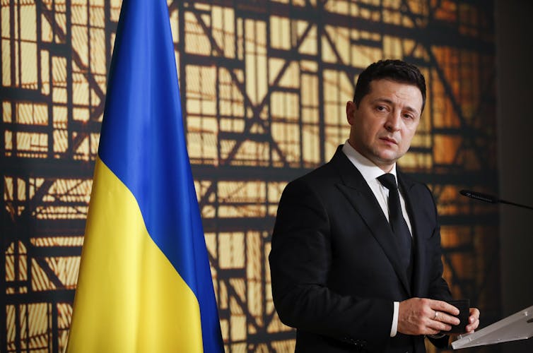 Ukrainian president Volodymyr Zelensky stands at a podium looking solemn, in front of a Ukrainian flag
