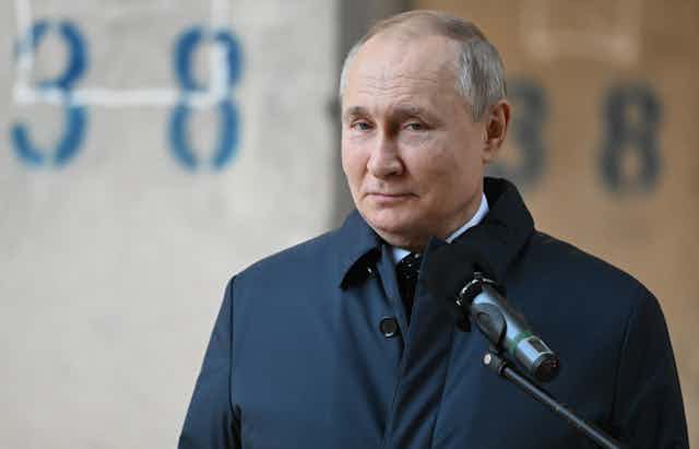 Vladimir Putin wearing a dark blue coat and standing behind a microphone