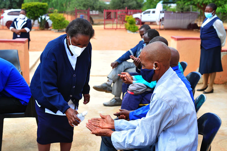 A row of people sitting in chairs outdoors while a woman dispenses hand sanitizer