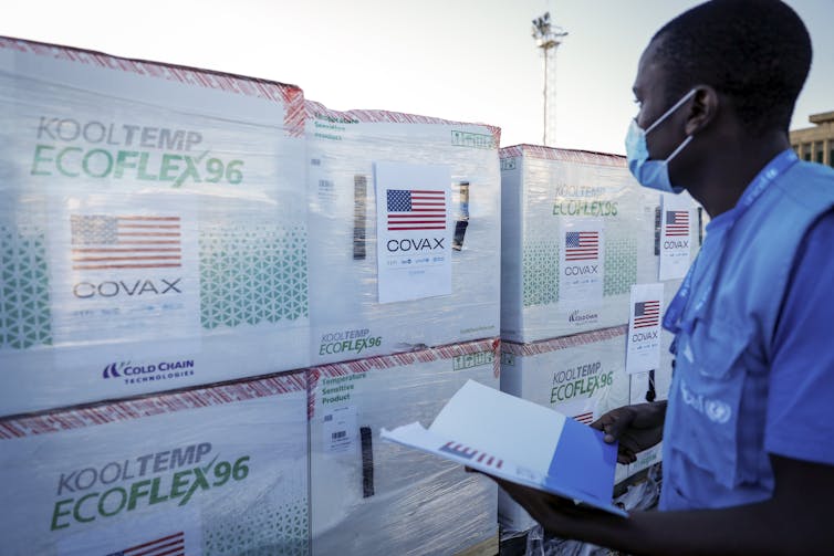 A worker wearing a surgical mask surveys a stack of boxes with the sign COVAX affixed to them