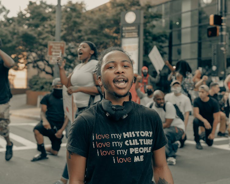 A young Black man is seen in a T-shirt reading 'I love my history, I love my culture, I love me.'