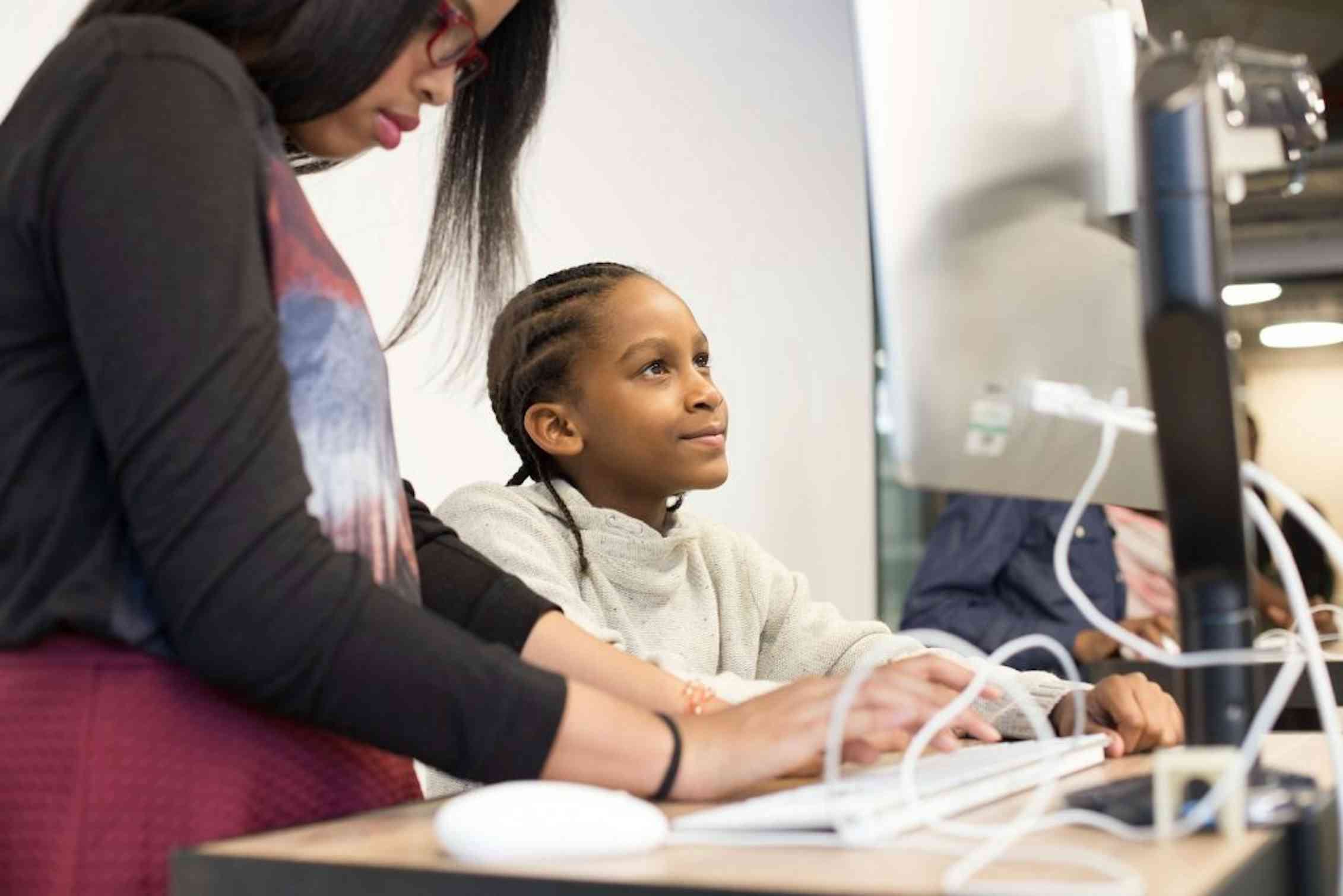 A Black boy works at a computer assisted by a Black teacher.