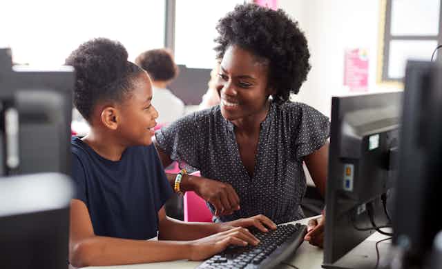 A Black youth is seen at a computer smiling at a Black teacher assisting her at a keyboard