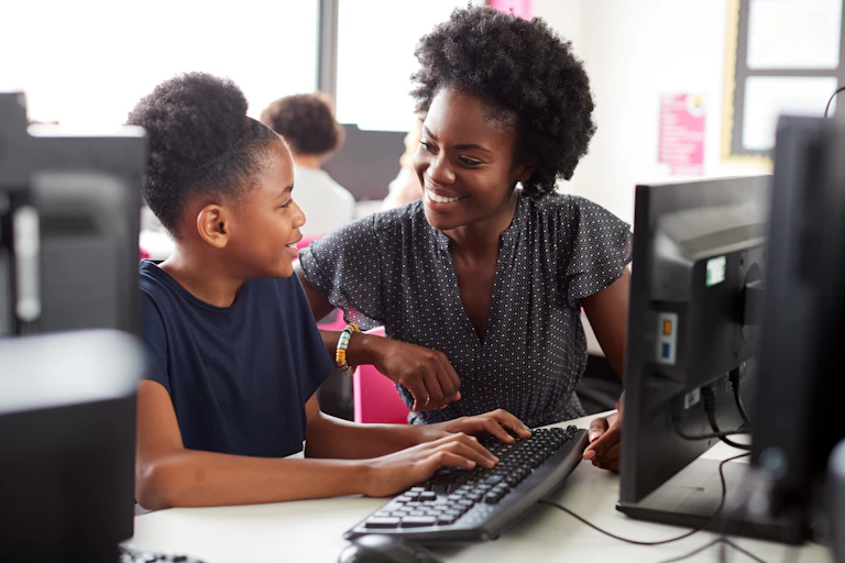 A Black youth is seen at a computer smiling at a Black teacher assisting her at a keyboard