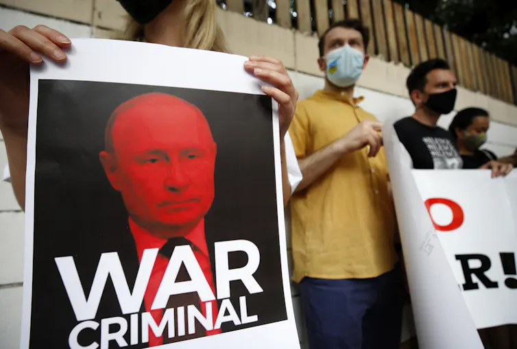 Primer plano de alguien sosteniendo un cartel con una foto de Putin en rojo, cubierto con un gran texto en blanco que dice 'criminal de guerra'