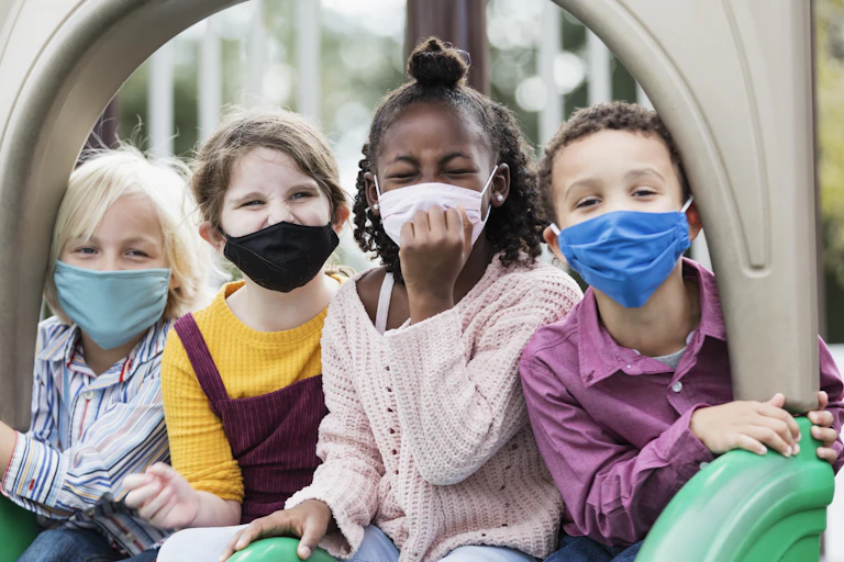 Four chldren wearing masks sit together on playground equipment.