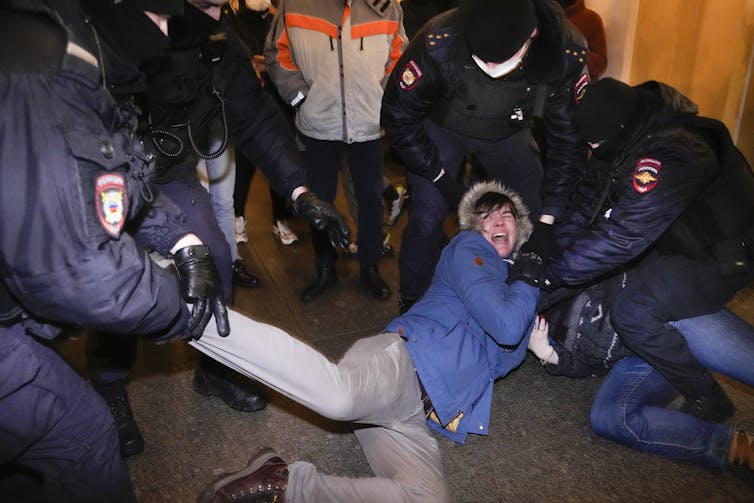 A woman in a blue jacket and grey pants lies on the ground crying out as police restrain her.