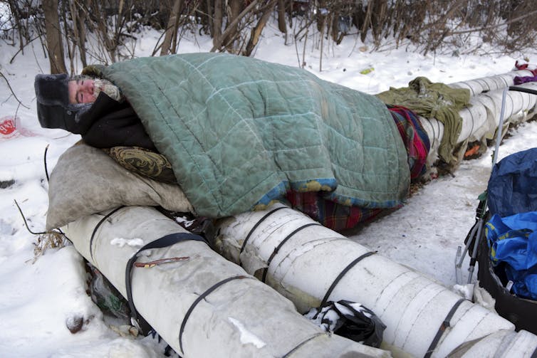 A bearded man in a black hat man lies under a stained blue blanket and on top of large pipes.