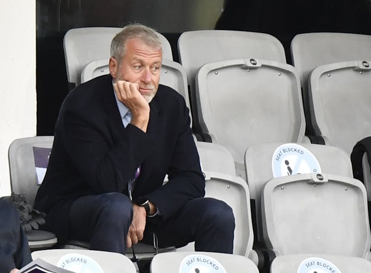 A grey-haired man in a navy jacket sits in a sports stadium with his hand under his chin.