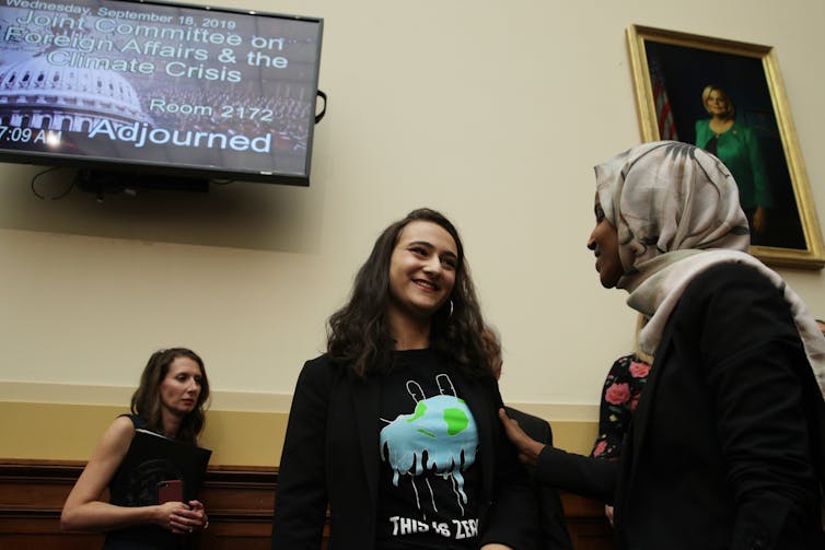 A woman speaks with a U.S. House member with a TV screening showing a hearing name behind them.