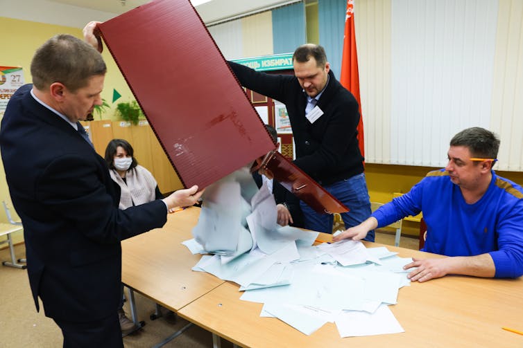 Four people sit and stand around a table and dump out white ballots of paper from a large maroon envelope.
