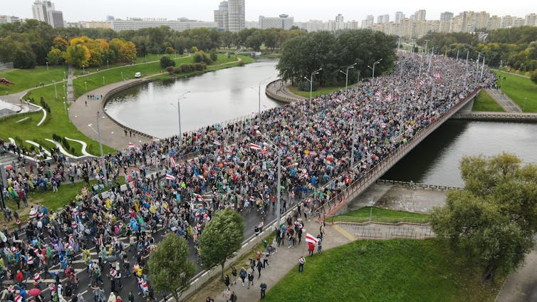 An aerial shot shows a large crowd of people marching across a bridge over a river on a grey day