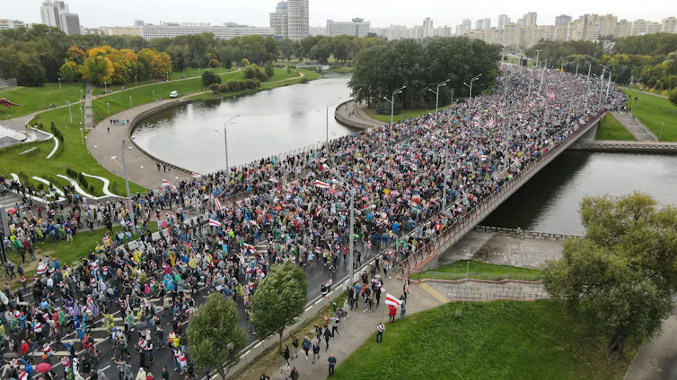 An aerial shot shows a large crowd of people marching across a bridge over a river on a grey day