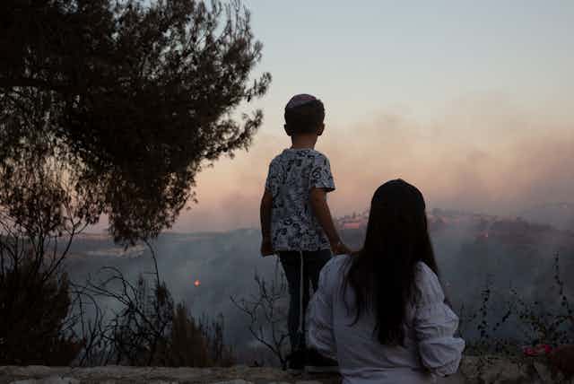 A boy stands on a wall with his mother behind him looking at fires blazing on hills with smoke wafting in front of them. 