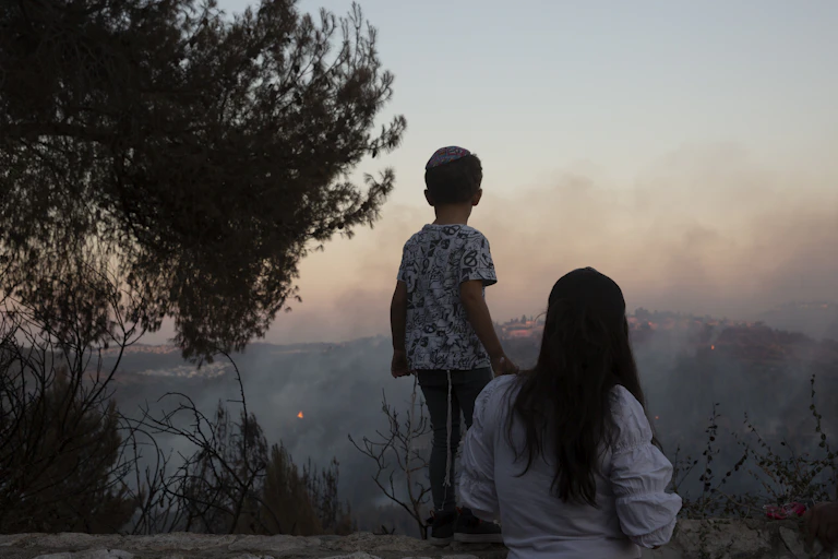 A boy stands on a wall with his mother behind him looking at fires blazing on hills with smoke wafting in front of them.