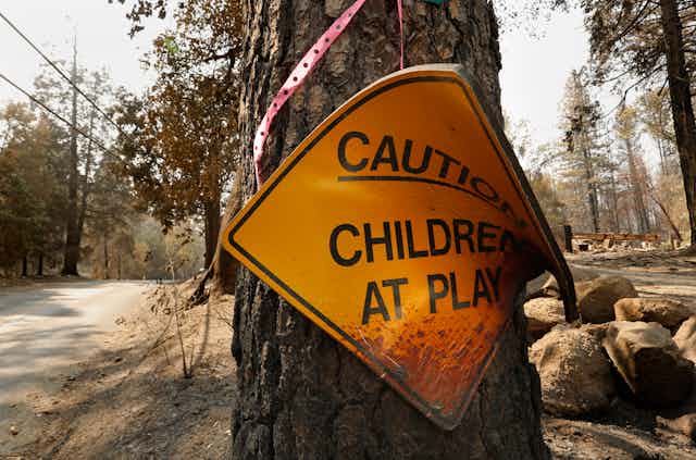 The diamond-shaped sign is attached to burned tree and curled over with burn marks and burned ground behind it.