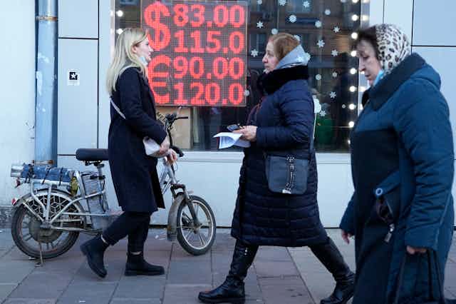 Two women in head kerchiefs and a blonde woman pushing a bicycle walk past a currency exchange display screen.