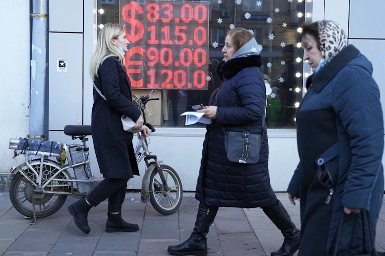 Two women in head kerchiefs and a blonde woman pushing a bicycle walk past a currency exchange display screen.