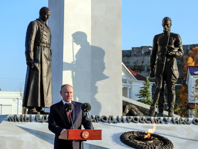 A bald man stands at a podium with statues of two men and an eternal flame behind him.