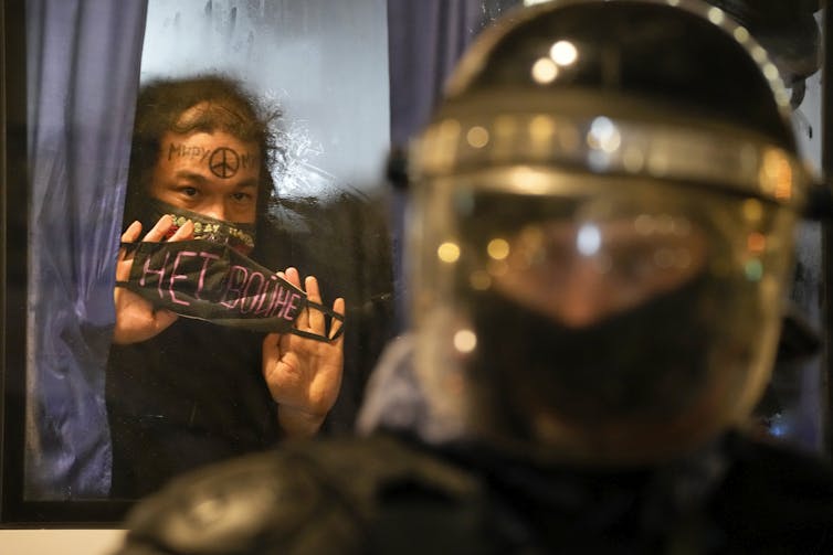 A person with a peace sign painted on their forehead holds up a sign against the glass of a police bus that says 'no war' with an armoured police officer in the foreground.
