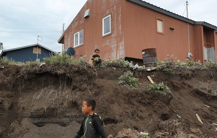 Two children play on an eroding hill with a house now at the edge of an eroded cliff that stands about one-story high.