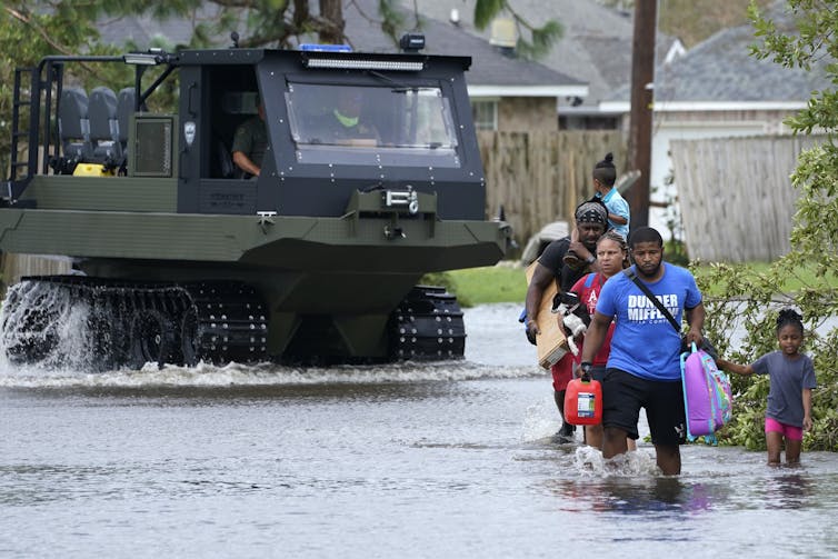 Three adults and two young children wade through flood water carrying suitcases.