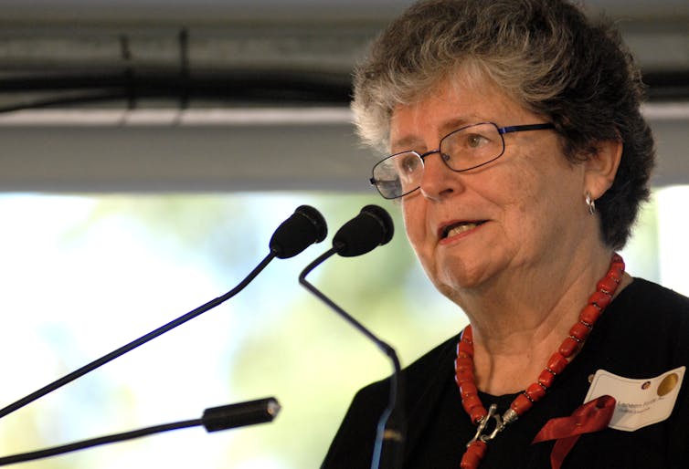 older woman speaking at a microphone, wearing glasses