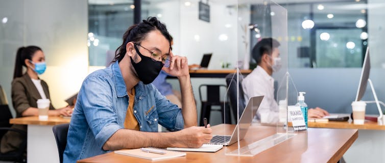 Man in a mask sits at his work desk, next to his female colleagues.