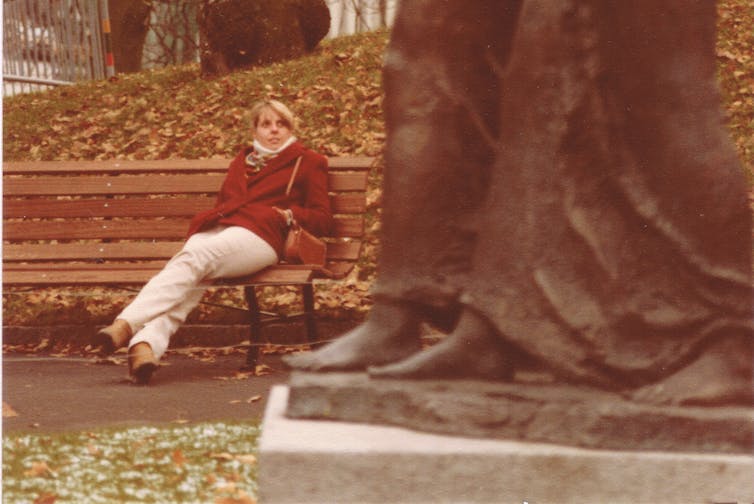 woman sits on bench, wrapped in coat and scarf, and stares at statue