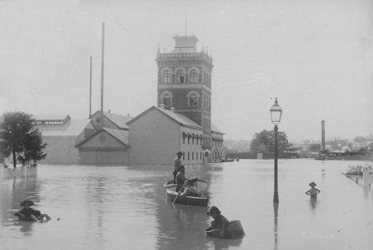 Black and white image of men in a small boat in front of a tall brick building that is half submerged in flood water. The building had 'West End Brewery' written on it