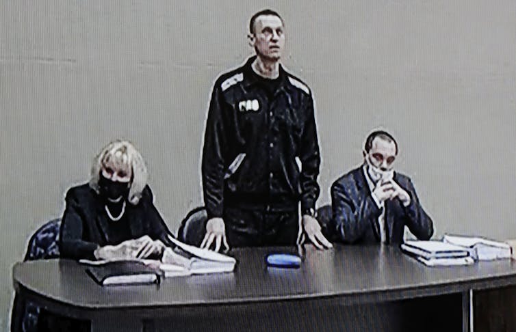 Russian opposition leader Alexei Navalany seen in a court video. In the low quality screenshot, he is standing at a table with two people sitting on either side of him.