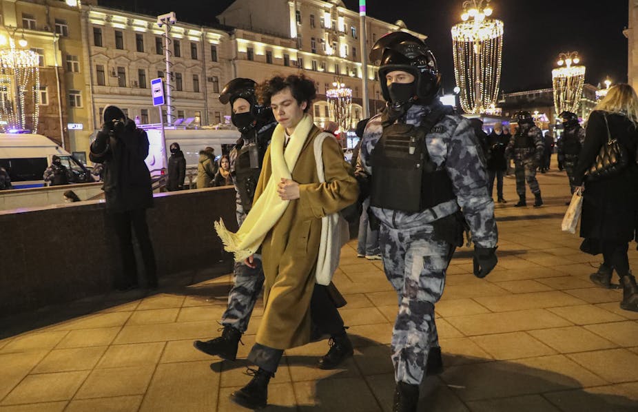 A young person in a long coat and scarf being walked by two police officers in heavy riot gear