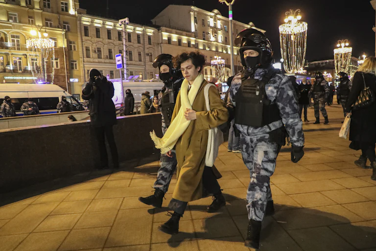 A young person in a long coat and scarf being walked by two police officers in heavy riot gear