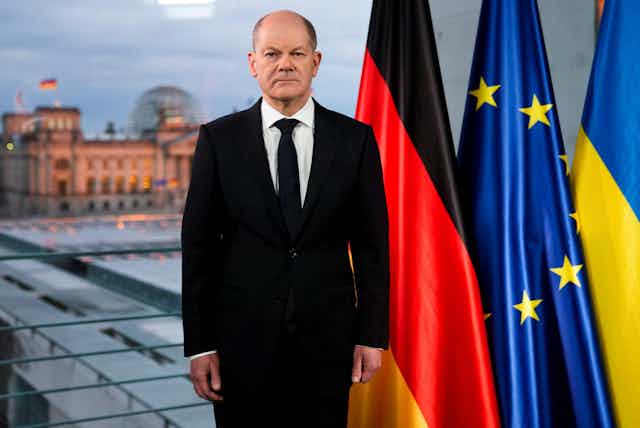 Olaf Scholz standing in front of the German, EU and Ukranian flags.