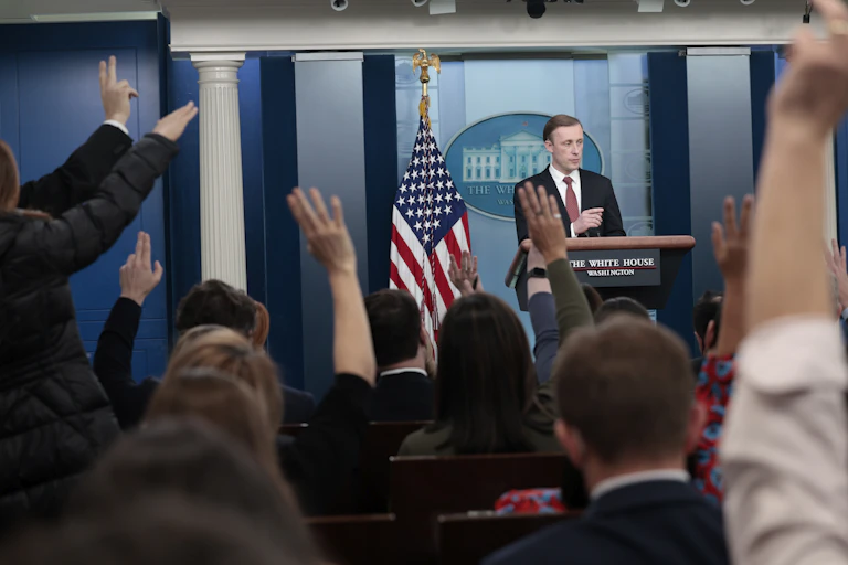 A man in a suit at a lectern that says "The White House" on it, answering questions from many people with raised hands.