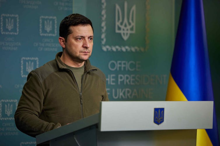 A man in a green sweater behind a lectern with the blue-and-yellow Ukrainian flag in the background.