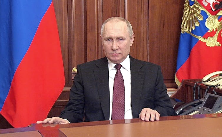 A balding man in front of two flags sitting at a desk in a dark suit, white shirt and red tie, looking serious.