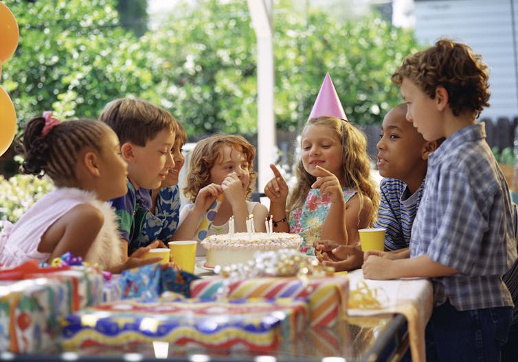Young kids gathered around a birthday cake at a table, with birthday girl preparing to blow out candles.