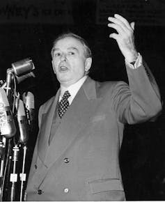 A black-and-white photo shows a man in a suit and tie gesturing as he speaks into a microphone.