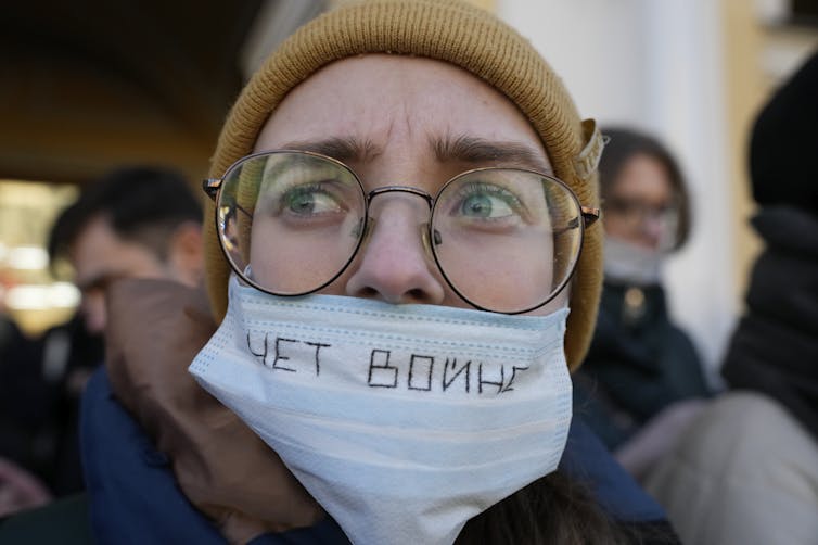A person wearing a hat, large eyeglasses and a surgical mask with Cyrillic handwriting on it