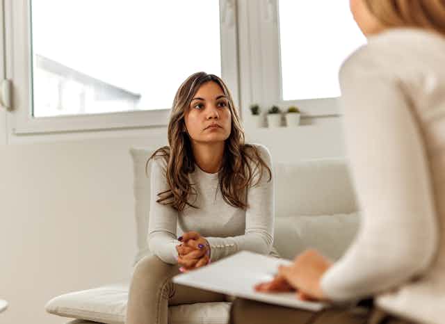 A seated woman with her hands folded, looking at another woman holding a clipboard, seen from behind.
