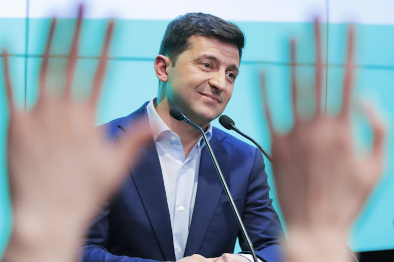 A man in a blue jacket and microphones in front of him smiles as people raise their hands to ask him questions.